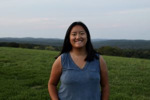 Mia Winslow smiling, in a blue-gray tank top, standing in front of a grassy expanse with mountains and a sunset in the background